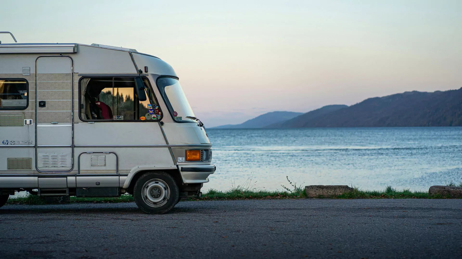 A white camper van, equipped with an RV surge protector, is parked by a calm lake with mountains visible in the distance under a clear sky.