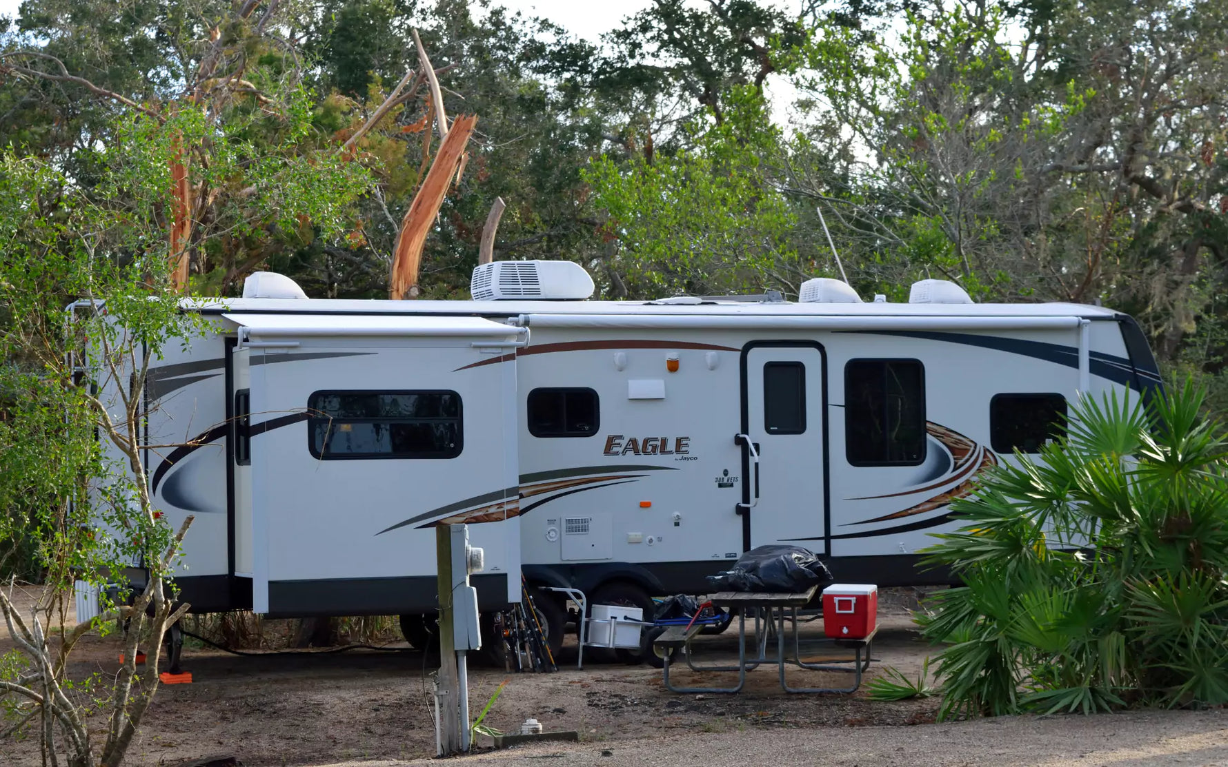 A travel trailer parked at a campsite, with a red portable refrigerator visible beside it.