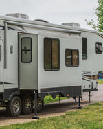 A light gray and black travel trailer parked outdoors on grass and dirt, with the side door open and support legs extended.