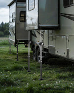 A beige and gray travel trailer parked on grass with its support legs extended outdoors.
